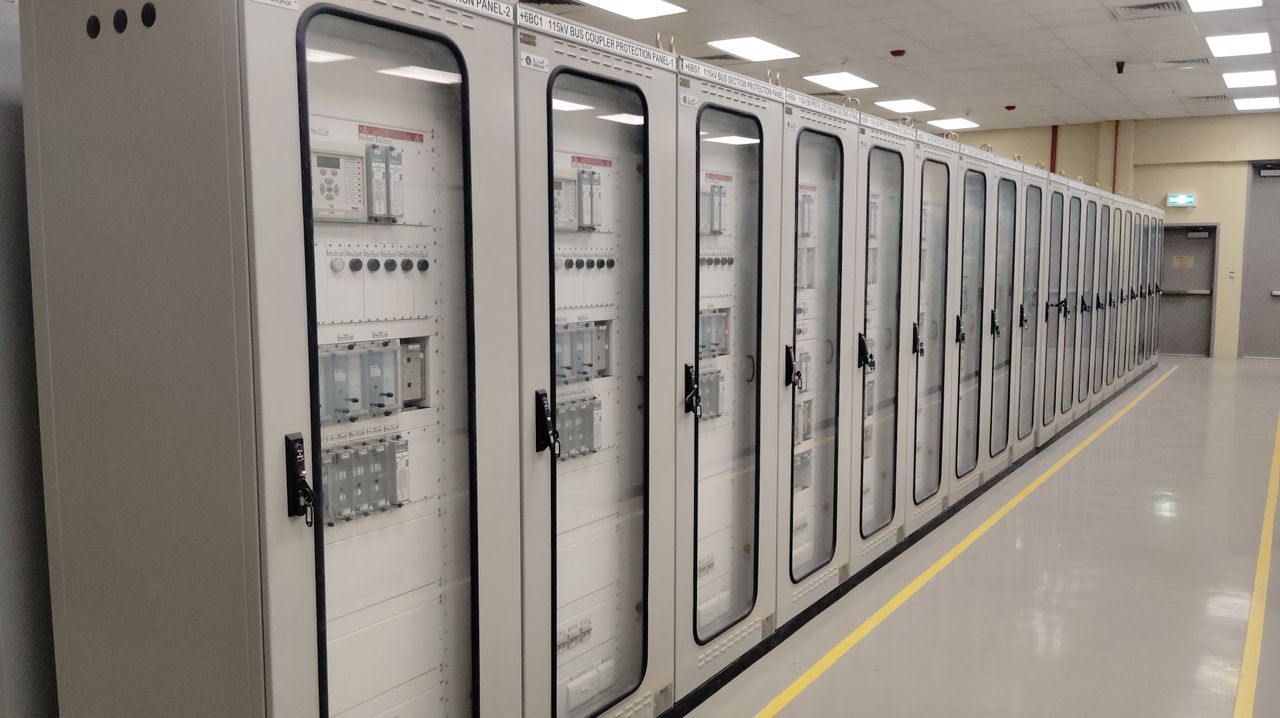 View of an industrial control room featuring rows of electrical panels in a well-lit environment.